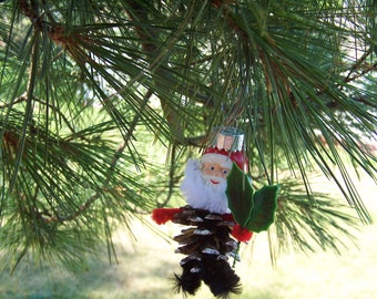 May include: A handmade Christmas ornament featuring a Santa Claus figure with a white beard and red hat. The figure is made from a pine cone and has a green holly leaf attached to it.