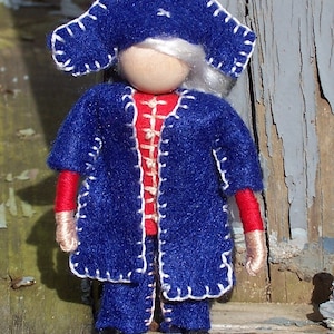 May include: A felt doll wearing a blue hat with white stitching and a blue coat with white stitching. The doll has a red and white striped shirt and a wooden head.