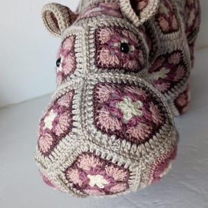 May include: A handmade crochet hippo in shades of gray, pink, and white. The hippo is crafted with a granny square pattern, featuring black button eyes and small, rounded ears. The plush toy is designed to resemble a hippo.