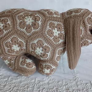 May include: A handmade crochet bunny in shades of brown and white. The bunny is made with a floral pattern and has long ears and black eyes. This soft sculpture is a decorative item.