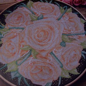 May include: A close-up of a cross-stitch embroidery of a bouquet of peach-colored roses with green leaves on a black background with white dots.
