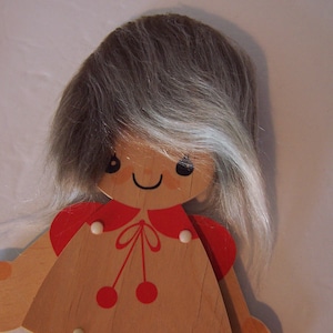 May include: Wooden doll with a smiling face, gray hair, and a red dress. The doll has a simple design with a cheerful expression. The hair is made of a soft, textured material. The red dress has white accents and cherry-like details.
