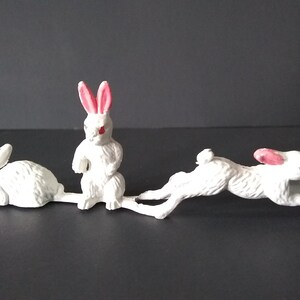 May include: Three white plastic toy rabbits with pink ears and red eyes. The rabbits are in different poses, one is lying down, one is standing up, and one is jumping.