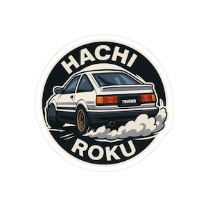May include: A sticker featuring a white and black sports car with the text "TRUENO" on the back. The car is drifting, with smoke billowing from the tires. The sticker has the words "HACHI ROKU" in a circular design.