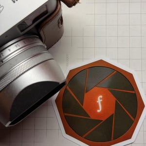May include: A silver Fujifilm X100V camera with a brown leather case sits next to a sticker. The sticker features a stylized camera aperture design in brown, black, and orange, with a white "f" in the center.