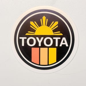 May include: A circular sticker featuring a black background with a yellow sun icon at the top, the white text TOYOTA in the center, and three vertical rectangular stripes in shades of orange, light orange, and yellow at the bottom.