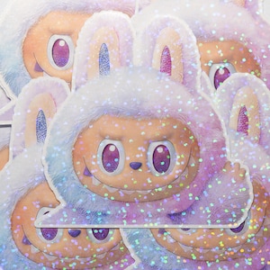 May include: A collection of stickers featuring a cartoon character with a furry, bunny-like hood in shades of purple, blue, and peach. The character has large purple eyes, a wide grin, and a glittery, iridescent finish.
