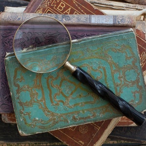 K&ouml;nnte beinhalten: Eine Vintage-Lupe mit schwarzem Griff liegt auf einem Stapel antiker B&uuml;cher. Die B&uuml;cher haben abgenutzte Einb&auml;nde in T&uuml;rkis-, Braun- und Lilat&ouml;nen mit goldgepr&auml;gten Designs. Die Lupe hat einen Messingrahmen.