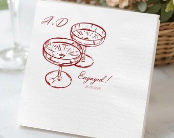 May include: White cocktail napkins featuring a red sketch of two champagne glasses. The text "A.D" is above the glasses, and "Engaged!" with a date is below. The napkins are stacked on a white surface.