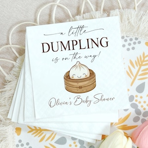 May include: White paper napkins with the text "a little DUMPLING is on the way!" and "Olivia's Baby Shower". The napkins feature a cartoon dumpling in a bamboo steamer. Baby shower theme.