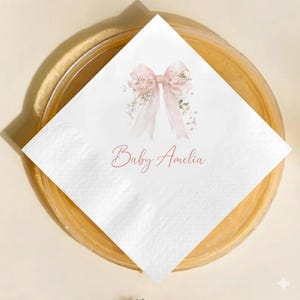 May include: White paper napkin with a watercolor design of a pink bow and floral accents. The name "Baby Amelia" is written in a delicate script below the bow. The napkin is placed on a gold-toned plate.