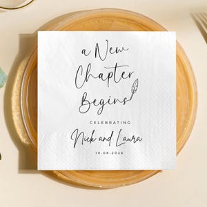 May include: A white square napkin with the text "a New Chapter Begins" in elegant script, celebrating "Nick and Laura" with the date 10.08.2026. The napkin rests on a clear plate.