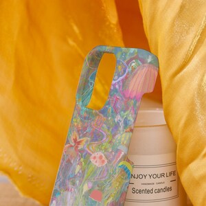 May include: A colorful phone case with a vibrant jellyfish design in shades of pink, blue, and yellow. The case is held in front of a white candle with the text "NJOY YOUR LIFE" and "Scented candles". The background is a bright yellow fabric.