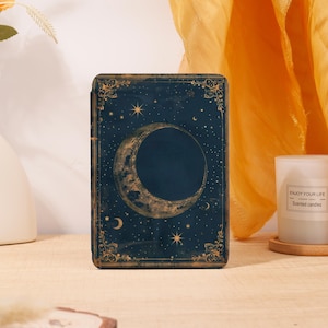 May include: A dark blue, rectangular box with a celestial design featuring a crescent moon, stars, and decorative borders. The box has a vintage aesthetic, with gold accents and a textured appearance. The box is likely for storing small items.