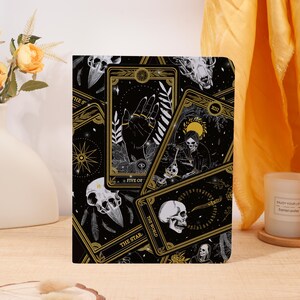 May include: A black journal featuring a tarot card design with gold accents. The cover displays various tarot cards with illustrations of skulls, hands, and celestial elements. The words "FIVE OF" and "THE STAR" are visible.