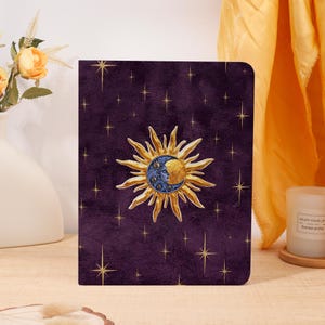 May include: A rectangular notebook with a deep purple cover featuring a central gold sun and blue moon illustration surrounded by scattered gold stars. The cover has a textured appearance and rounded corners, showcasing a celestial design.