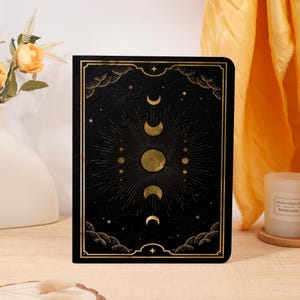 Könnte beinhalten: Schwarzes Notizbuch mit goldenem Himmelsdesign mit Mondphasen und strahlendem Sonnenaufgang. Das Cover hat einen goldenen Rand mit dekorativen Wolken- und Sternakzenten. Das Notizbuch wird auf einer hellen Oberfläche präsentiert.