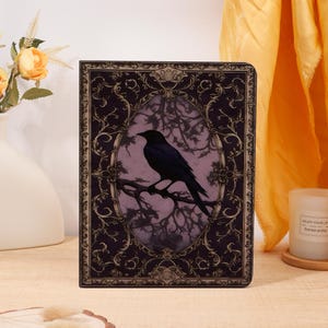 May include: A decorative book cover featuring a black raven perched on a branch within an oval frame. The frame is set against a purple background with a dark purple border and ornate gold filigree. The overall design has a gothic or vintage aesthetic.