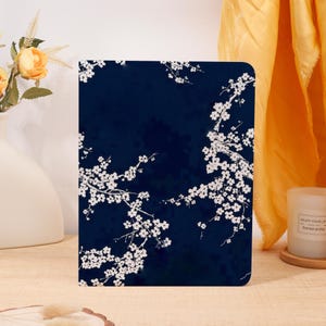 Könnte beinhalten: Ein dunkelblauer Notizbuchumschlag mit einem weißen Blumendesign. Das Design zeigt Zweige mit weißen Blüten. Das Notizbuch ist rechteckig und hat abgerundete Ecken. Der Hintergrund ist einfarbig dunkelblau.