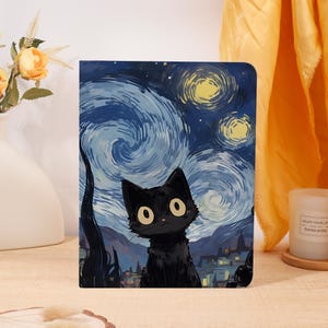 May include: A rectangular notebook featuring a black cat illustration set against a swirling blue and yellow night sky background. The artwork depicts a starry night scene with a dark silhouette of a town at the bottom and a large, expressive black cat.