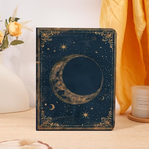 May include: A dark blue journal with a celestial design featuring a crescent moon and stars. The cover has a gold border with decorative flourishes. The journal is a hardcover and appears to be new.