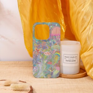 May include: A colorful phone case featuring a vibrant underwater scene with jellyfish, flowers, and mushrooms. A white candle with the text "ENJOY YOUR LIFE" sits next to the phone case on a wooden surface. The background is a golden yellow fabric.