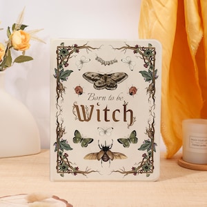 May include: A rectangular notebook with a cream-coloured cover featuring the text Born to be Witch in brown lettering. The design includes illustrations of various moths, butterflies, a beetle, a caterpillar, and two ladybirds framed by brown vines.