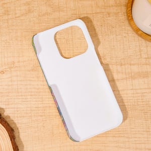 May include: White phone case with a floral design on the edges. The case has a cutout for the camera and buttons. The case is laying on a wooden surface.