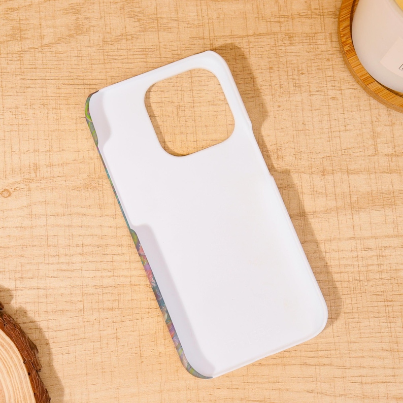 May include: White phone case with a floral design on the edges. The case has a cutout for the camera and buttons. The case is laying on a wooden surface.