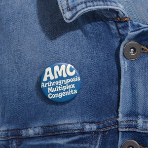 May include: A blue button pin with the letters "AMC" in white, above the words "Arthrogryposis Multiplex Congenita" in white. The pin is attached to a blue denim jacket.
