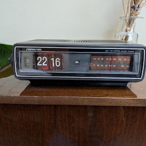May include: A vintage black Soundesign AM-FM digital clock radio. The clock displays the time 22:16 in red digits. The radio dial is visible, and the unit sits on a wooden surface. A small glass vase with dried flowers is in the background.