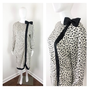 May include: A white and black patterned dress with a black satin bow at the neckline. The dress has long sleeves and a fitted silhouette.