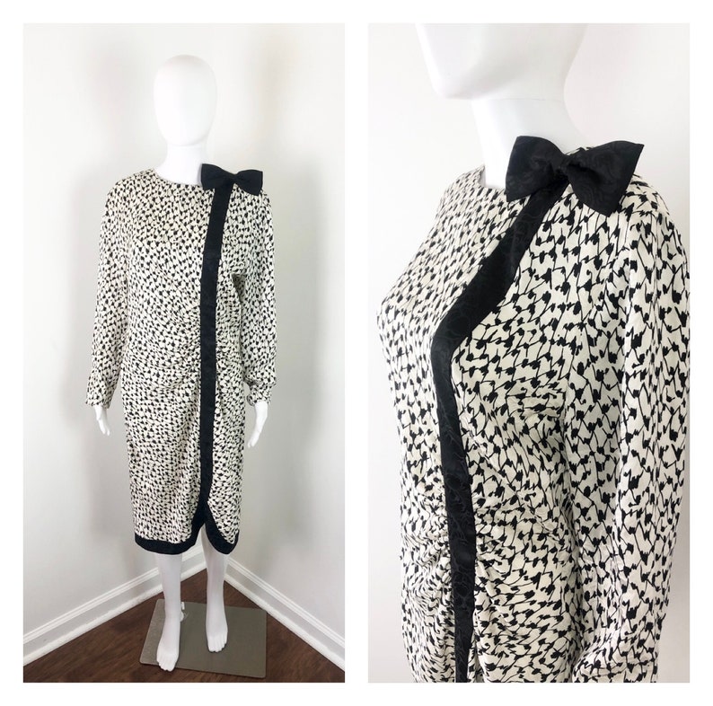 May include: A white and black patterned dress with a black satin bow at the neckline. The dress has long sleeves and a fitted silhouette.