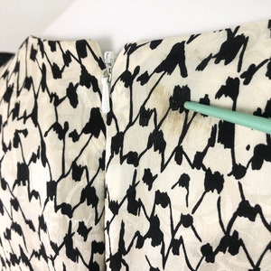 May include: Back view of a white and black patterned dress with a zipper closure. The dress has a subtle sheen and a geometric pattern.
