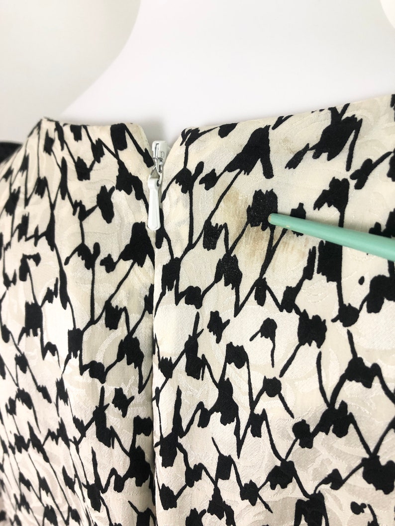May include: Back view of a white and black patterned dress with a zipper closure. The dress has a subtle sheen and a geometric pattern.