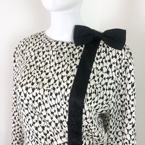 May include: A white and black patterned blouse with a black satin bow on the shoulder.