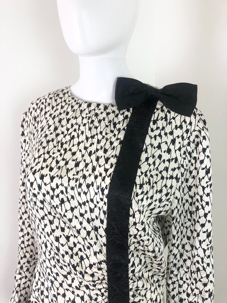 May include: A white and black patterned blouse with a black satin bow on the shoulder.