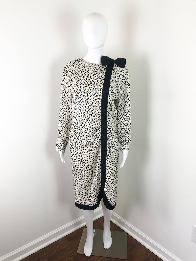 May include: A white dress with a black and white geometric print. The dress has a black bow at the neckline and a black trim around the hem.