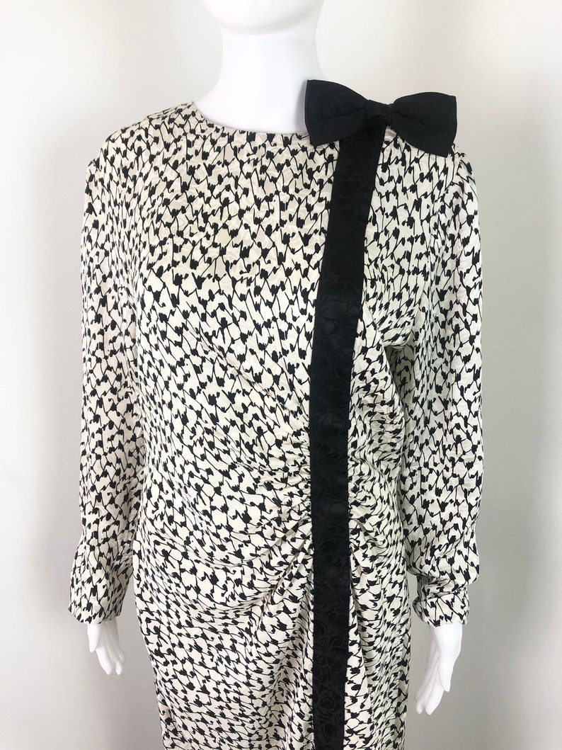 May include: A white and black patterned dress with a black bow at the shoulder. The dress has a long sleeve and a fitted silhouette.