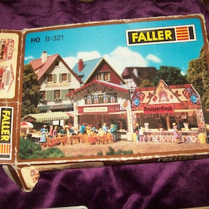 May include: A model train set box featuring a detailed miniature village scene with shops, restaurants, and people. The box is labeled "FALLER" and "HO B-321". The image shows a colorful depiction of a European town with blue skies.