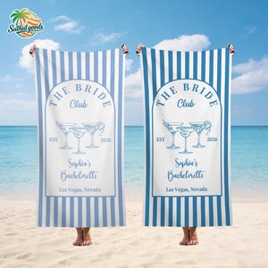 May include: Two blue and white striped beach towels with the text "THE BRIDE Club" and "Sophia's Bachelorette Las Vegas, Nevada" in a light blue font. The towels have a design of cocktail glasses. The towels are held up on a beach.