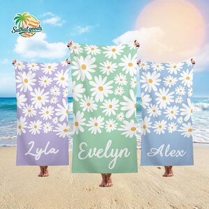 May include: Three beach towels with daisy patterns and personalized names. The towels are in shades of lavender, mint green, and light blue. Each towel has a white daisy pattern with a yellow center and a name at the bottom.