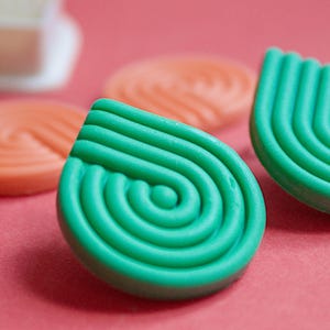 May include: Close-up of several soap bars in a teardrop shape with a spiral design. The front-most bar is bright green, while others are orange. The background is a solid pink color. The soap bars have a textured, ridged surface.