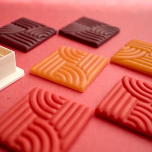 Könnte beinhalten: Eine Auswahl quadratischer, strukturierter Objekte in Rot-, Orange- und Brauntönen, angeordnet auf einer rosa Oberfläche. Die Objekte weisen ein sich wiederholendes geometrisches Muster auf. Ein weißes rechteckiges Objekt befindet sich im Hintergrund.