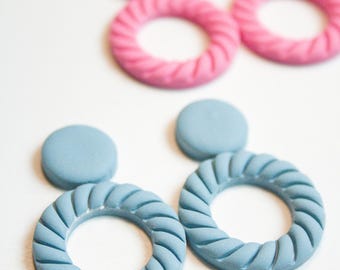 May include: Close-up of handmade earrings in two pairs. One pair is light blue, the other is pink. Each earring has a circular design with a textured, rope-like pattern. The earrings are on a white surface.