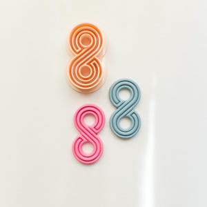 May include: Three number eight shaped cookie cutters in different colors: peach, pink, and blue. The cutters are made of plastic and are on a white background. The number eight design is a continuous loop.