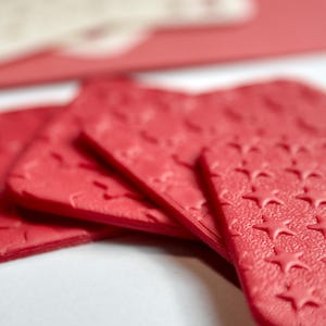 May include: Close-up of several red, rectangular, textured objects with a star pattern. The objects are arranged in a fan shape, with a blurred white and red background. The objects appear to be decorative or functional.