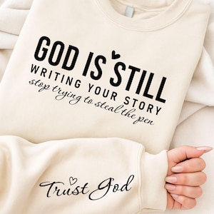 Puede incluir: Sudadera color crema con el texto negro "GOD IS STILL WRITING YOUR STORY stop trying to steal the pen". La manga presenta las palabras "Trust God" en una fuente cursiva. Una pequeña pegatina redonda en la esquina superior izquierda dice "FREE BONUS BUNDLE".