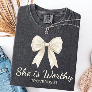 May include: Dark gray t-shirt with a cream-colored bow graphic and the text "She is Worthy PROVERBS 31". A silver necklace with a pendant is on the shirt. The shirt is folded next to a pair of blue jeans.