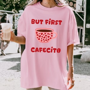 May include: Pale pink t-shirt featuring the words "BUT FIRST CAFECITO" in red. The design includes a coffee cup filled with brown liquid and decorated with red hearts. The shirt has a relaxed, oversized fit.